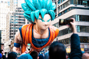 Goku gigante sendo carregado por várias pessoas em uma cidade grande.