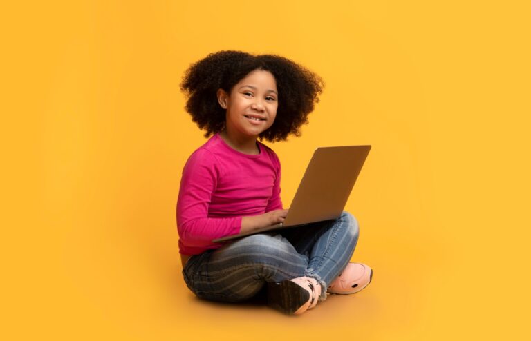 Criança negra, de cabelos cachedos, sorrindo segurando um laptop olhando para a câmera.